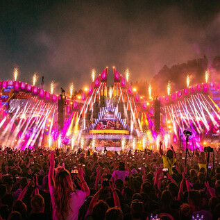 A lively music festival with a huge stage surrounded by colorful lights and fireworks. The crowd cheers and is full of energy. | © Geoffrey Hubbel
