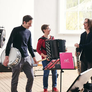 Tři hudebníci stojí v světlé místnosti a hrají na různé nástroje. Jeden hraje na akordeon, zatímco ostatní drží buben a dřevěný dechový nástroj. | © Andreas Hechenberger