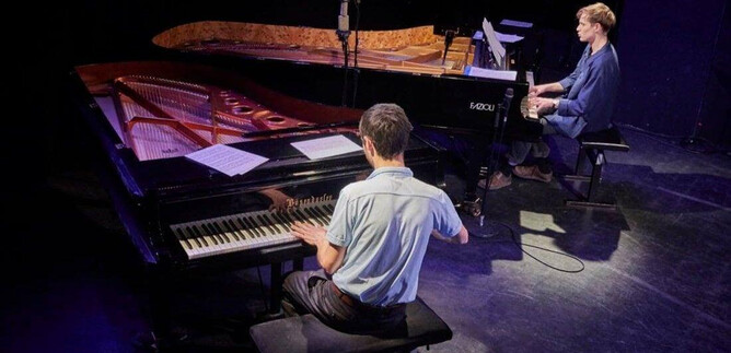 Two pianists are sitting on stage and playing together. One pianist is at a piano, while the other is seated at an additional grand piano. | © Jazzit