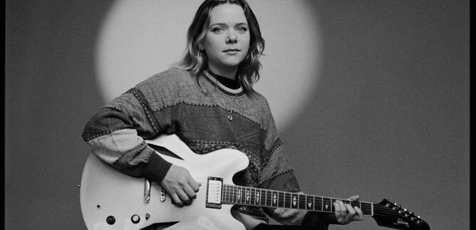 Eine Person sitzt mit einer Gitarre in der Hand. Sie trägt einen Pullover mit grafischem Muster und schaut direkt in die Kamera. | © Jazzit_Ella Zirina