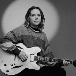 Une personne est assise avec une guitare à la main. Elle porte un pull à motif graphique et regarde directement la caméra. | © Jazzit_Ella Zirina