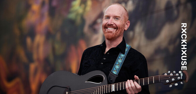 Ein lächelnder Mann hält eine Gitarre in der Hand. Im Hintergrund sind farbige, abstrakte Muster zu sehen. | © Lisa Sciliano