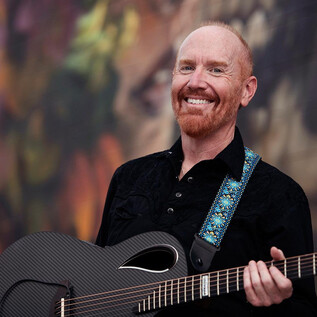 Egy mosolygó férfi gitárt tart a kezében. A háttérben színes, absztrakt minták láthatók. | © Lisa Sciliano