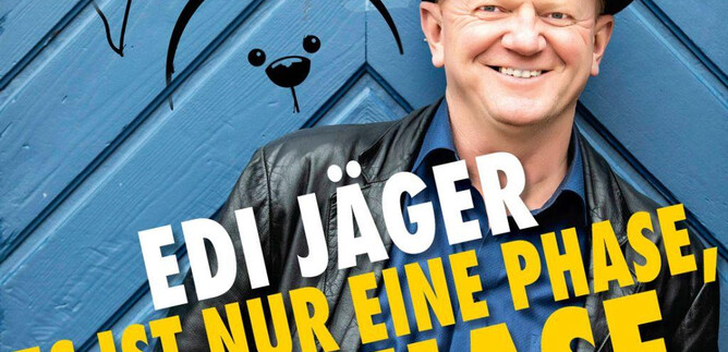 A smiling man is wearing a black hat and a black jacket in front of a blue wall. In the background, cartoon-like rabbit illustrations can be seen. | © Kurt-Sixtl_Neuhofen