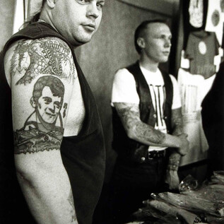 Two men with tattoos are standing at a table full of T-shirts. In the foreground, there is a man with a striking tattoo on his arm. | © Ruth Bayer