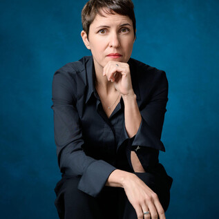 Osoba, která působí zamyšleně, sedí na stoličce s krátkými vlasy. Pozadí je modré a vytváří silný kontrast k tmavému oblečení. | © Christian Palm-Half