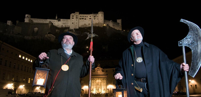Dva muži v historických oděvech stojí v noci s lampami před osvětleným hradem. Drží hůl a velkou sekeru a vážně se dívají do dálky. | © Reischenböck & Knoll