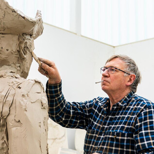 Un hombre mayor trabaja en una escultura de arcilla en un taller luminoso. Lleva una camisa de cuadros y sostiene una herramienta en la mano. | © Stephan Balkenhol/Bildrecht, Wien 2026/Noah Kronreif