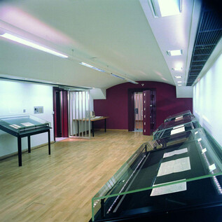 A modern exhibition space with glass display cases and a bright wooden floor. The walls are partially painted red, and there are various doors in the room. | © ISM