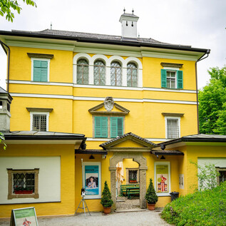 Un impresionante edificio amarillo con persianas verdes y una hermosa entrada. Rodeado de un jardín verde, irradia un carácter encantador y acogedor. | © Melanie Wressnigg
