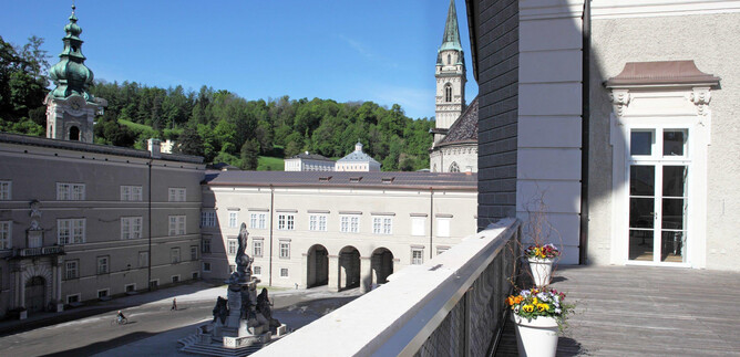 Una vista meravigliosa su una piazza storica con edifici e una fontana. Sullo sfondo ci sono colline verdi e il cielo è sereno e blu. | © DomQuartier