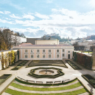 Un hermoso jardín con setos simétricos y una fuente central. En el fondo se pueden ver edificios y un cielo claro y azul. | © Salzburg Museum/Tolga Salpat