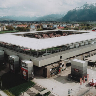 Moderní fotbalový stadion s prostornou tribunou. V pozadí jsou vidět hory a městská krajina.