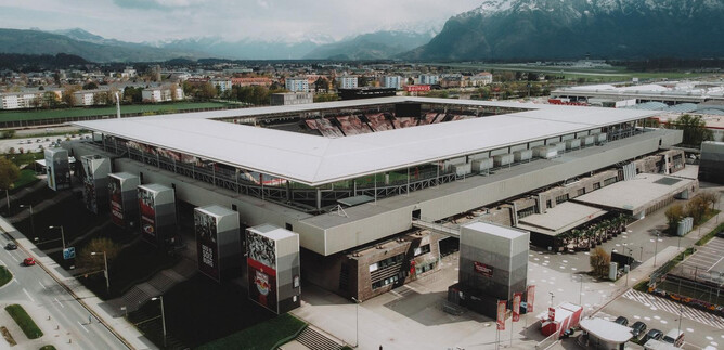 A modern football stadium with a spacious grandstand. In the background, mountains and an urban landscape can be seen.