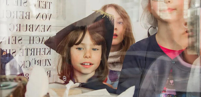 Un grupo de niños mira curiosamente a través de una ventana. Están vestidos con ropa festiva y observan objetos expuestos en una mesa. | © Eva trifft