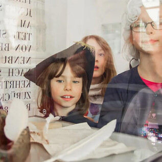 Un gruppo di bambini guarda curiosamente attraverso una finestra. Sono vestiti in abiti festivi e osservano oggetti esposti su un tavolo. | © Eva trifft