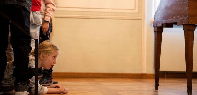 A girl is lying on the floor, looking curiously under a table. In the background, several people can be seen, also looking with interest. | © Erika Mayer