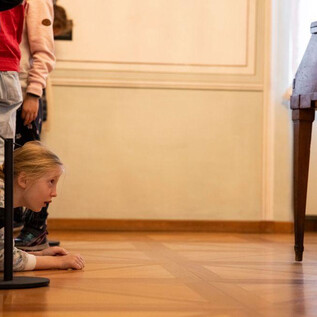 A girl is lying on the floor, looking curiously under a table. In the background, several people can be seen, also looking with interest. | © Erika Mayer