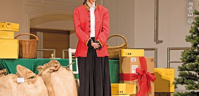 A cheerful performer in a red jacket stands in front of a festively decorated background with gifts and a Christmas tree. There is a festive atmosphere. | © Erika Mayer