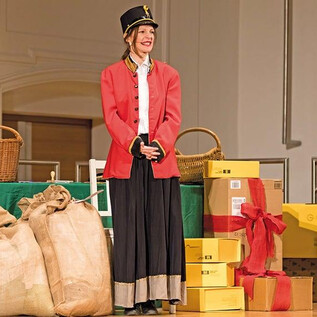 A cheerful performer in a red jacket stands in front of a festively decorated background with gifts and a Christmas tree. There is a festive atmosphere. | © Erika Mayer