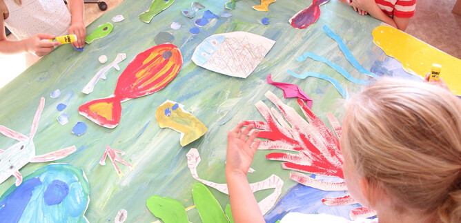 Dzieci malują żywy świat podwodny na dużym stole. Widać kolorowe ryby i rośliny z papieru. | © DomQuartier