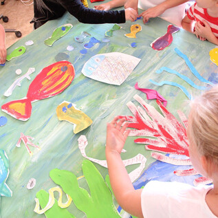 Gyerekek élénk víz alatti világot festenek egy nagy asztalon. Papírból készült színes halak és növények láthatók. | © DomQuartier