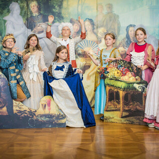 A group of children in historical costumes stands in front of a backdrop painting. They are happily posing and embodying various figures from the past. | © DomQuartier_Neumayr