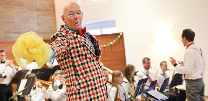 Ein fröhlicher Clown in einem karierten Anzug steht im Vordergrund und unterhält das Publikum. Im Hintergrund spielt ein Orchester mit verschiedenen Instrumenten. | © Franz Neumayr