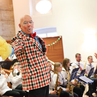 Un clown allegro in un completo a quadri è in primo piano e intrattiene il pubblico. Sullo sfondo suona un'orchestra con vari strumenti. | © Franz Neumayr