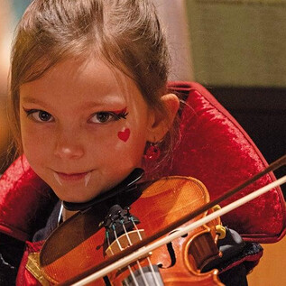 A small girl with a violin and a red cloak. She has a heart painted on her cheek and looks directly into the camera. | © Erika Mayer