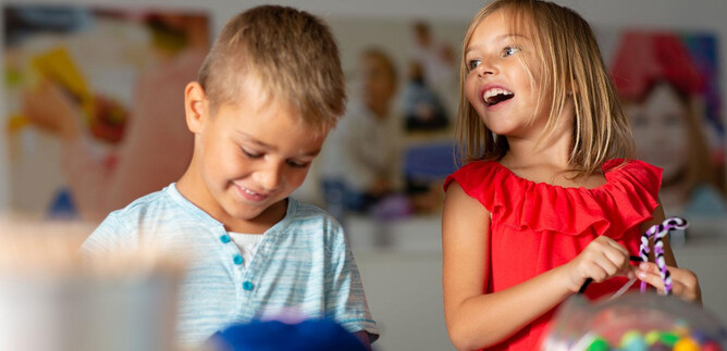 Dwoje dzieci bawi się i śmieje się razem. Chłopiec ma na sobie jasnoniebieską koszulę, a dziewczynka czerwoną sukienkę. | © Hannelore Kirchner