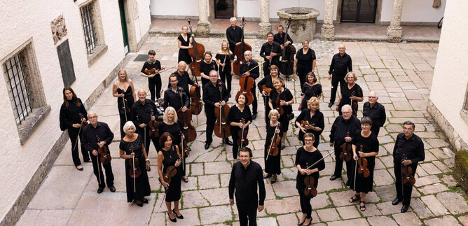 A large orchestra group is standing in the courtyard of a building. The musicians are wearing black clothing and holding their instruments. | © Salzburger Kulturvereinigung