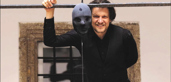 A man in black stands with a smile next to a hanging mask. In the background, an old wall and a window can be seen. | © Erika Mayer