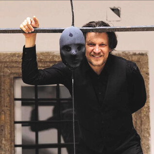 Un homme en noir se tient souriant à côté d'un masque suspendu. En arrière-plan, on peut voir un vieux mur et une fenêtre. | © Erika Mayer