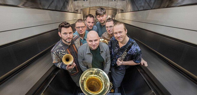 A group of seven musicians stands in a modern environment. They hold various wind instruments and pose on an escalator. | © Severin Koller