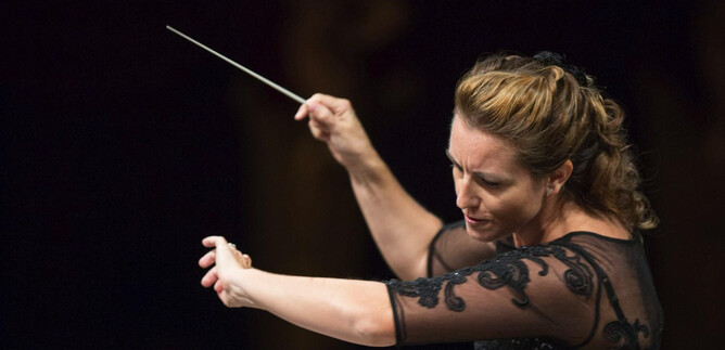 A conductor leads the orchestra with a baton. She has a focused expression and wears an elegant black dress. | © FSOA