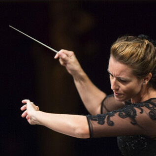 Eine Dirigentin führt das Orchester mit einem Taktstock. Sie hat eine konzentrierte Miene und trägt ein elegantes, schwarzes Kleid. | © FSOA