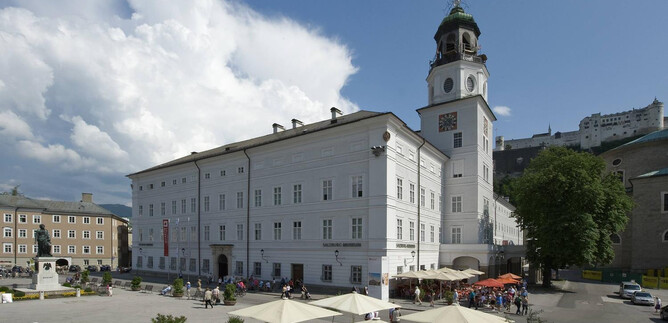 Ein historisches Gebäude mit einem markanten Turm in einer Stadt. Im Vordergrund sind Tische und Stühle unter parasolartigen Sonnenschirmen. | © Salzburg Museum