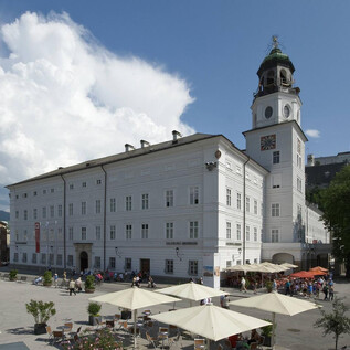 Historická budova s výraznou věží ve městě. V popředí jsou stoly a židle pod slunečníky podobnými parasolům. | © Salzburg Museum