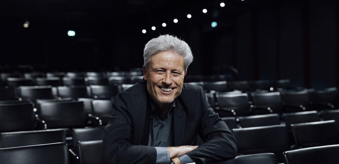 A smiling man in a suit sits in an empty theater. In the background, empty rows of seats are visible. | © Sky Sport Austria