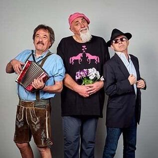 Drei Männer posieren vor einem neutralen Hintergrund. Der erste Mann spielt Akkordeon, der zweite trägt ein pinkes Outfit und der dritte wirkt elegant in Anzug und Sonnenbrille. | © Christian Streili