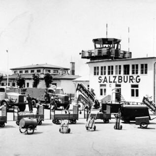 Egy történelmi fénykép a salzburgi repülőtérről, különböző járművekkel és egy irányítótoronnyal. A háttérben épületek és fák láthatók. | © Salzburger Flughafen GmbH