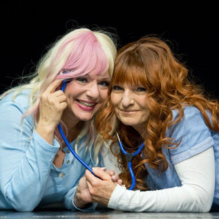 Two women in blue shirts are lying next to each other on stage. They are laughing and holding a stethoscope, indicating a medical or humorous performance. | © Christian Treweller