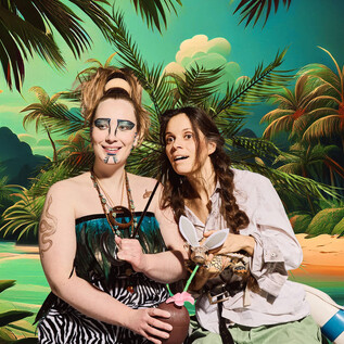 Two women are happily posing in front of a tropical background with palm trees. One woman is holding a coconut, while the other is showcasing animal accessories. | © Mathias Schuh