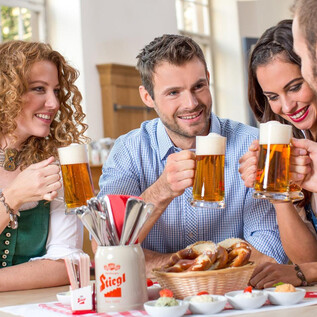 A group of four friends is sitting around a table, toasting with beer mugs. On the table lies a food basket with pretzels and snacks. | © Stieglbrauerei zu Salzburg GmbH