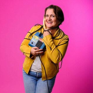 Eine fröhliche Frau hält ein blaues Telefon und lächelt. Der Hintergrund ist leuchtend pink. | © Stefan Joham