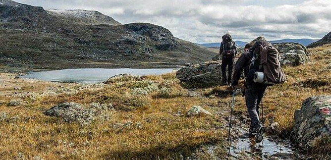 Zwei Wanderer ziehen durch ein grünes, hügeliges Gelände mit einigen Felsen und einem ruhigen See im Hintergrund. Der Himmel ist bewölkt, und die Landschaft wirkt wild und unberührt. | © Messezentrum Salzburg