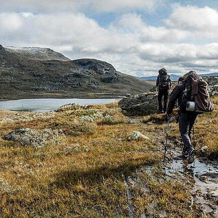 Zwei Wanderer ziehen durch ein grünes, hügeliges Gelände mit einigen Felsen und einem ruhigen See im Hintergrund. Der Himmel ist bewölkt, und die Landschaft wirkt wild und unberührt. | © Messezentrum Salzburg