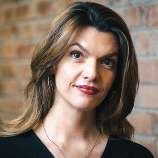 Egy hosszú, göndör hajú nő áll egy téglafal előtt. Barátságos arckifejezése van, és fekete felsőt visel. | © Ingo Pertramer