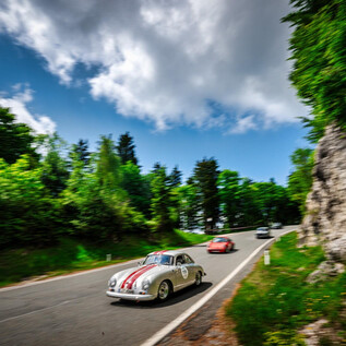 Ein klassisches Auto mit Rennstreifen fährt eine kurvenreiche Straße entlang. Umgeben von grünen Bäumen und einem blauen Himmel mit Wolken. | © motorsportpics.at